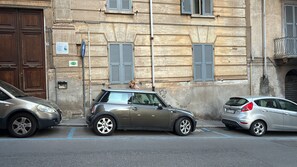 Straßenansicht mit parkenden Autos, einem Gebäude mit Fensterläden und einem Schild, das eine Bushaltestelle kennzeichnet.