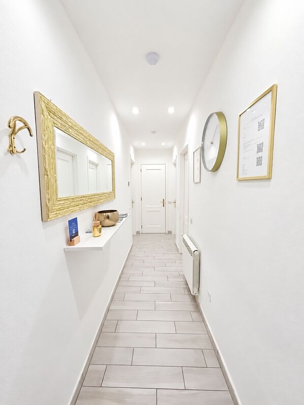 A hallway with a white floor, a wall-mounted mirror, a clock, and a framed document.