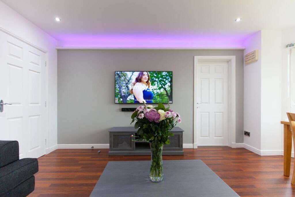 Un salon avec un téléviseur à écran plat fixé au mur, un canapé gris et un arrangement floral posé sur un tapis.
