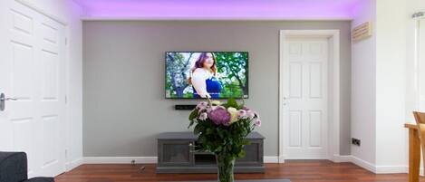 Un salon avec un téléviseur à écran plat fixé au mur, un canapé gris et un arrangement floral posé sur un tapis.
