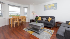 A modern living room with a grey sofa, wooden dining table, and chairs. There is a world map on the wall and a lamp on a side table.