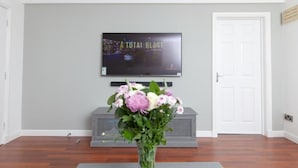 A living room with a flat-screen TV mounted on the wall, a grey entertainment unit below it, and a white door to the right.