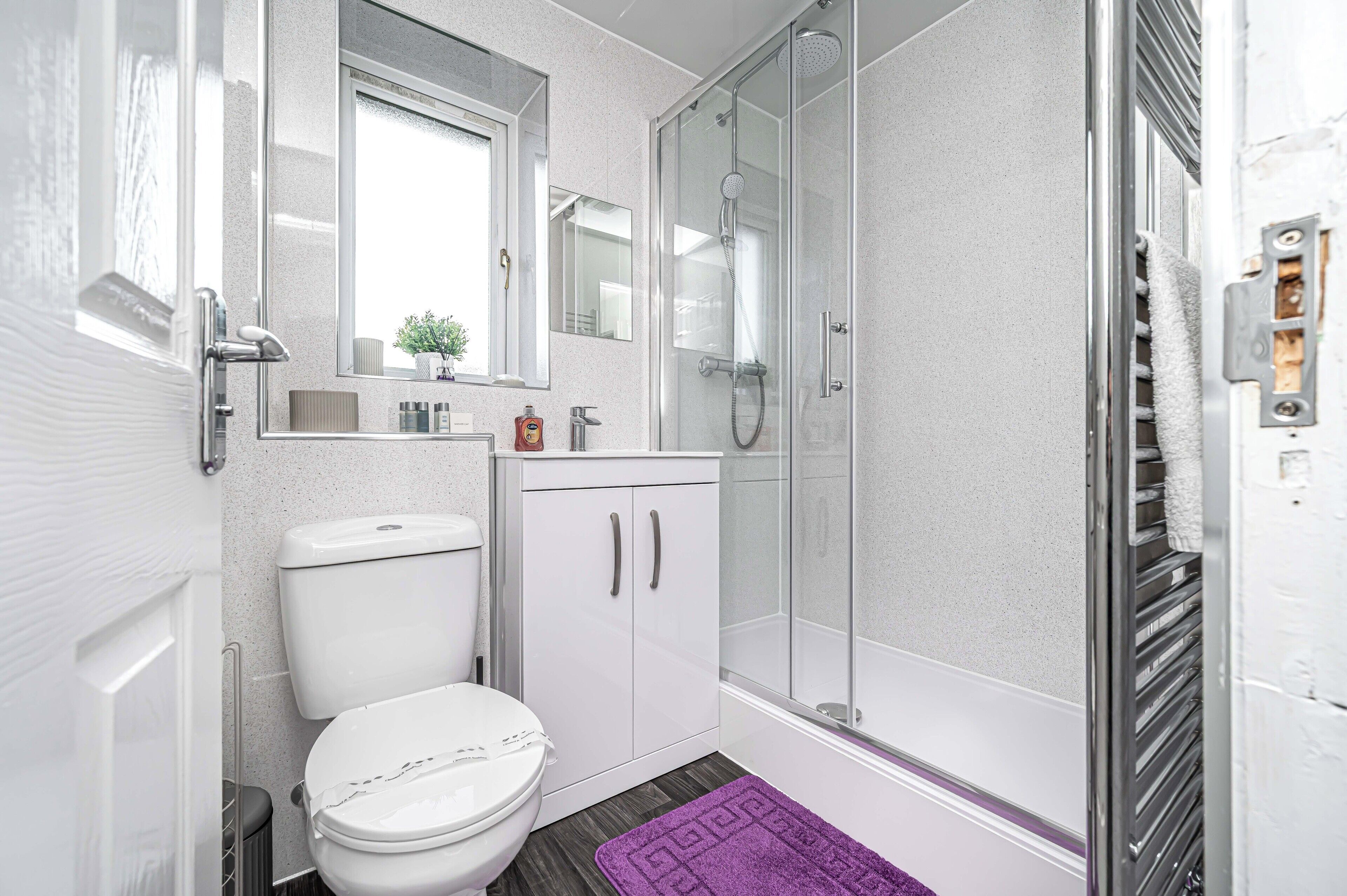 Une salle de bain comprenant des toilettes, un lavabo, un miroir, une douche avec des portes en verre et un tapis violet.