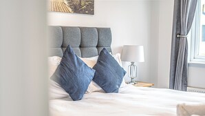 Un lit bien fait, avec une tête de lit rembourrée de couleur grise, du linge de lit blanc et deux oreillers bleus.