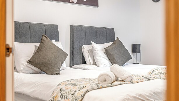 A neatly made bed with a grey headboard, white and grey pillows, and a rolled white towel. - Super Prime Train Station Inverkeithing by Three Bridges Apartments (Inverkeithing)