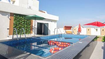 Una piscina en la azotea con un cielo azul despejado, una pared verde con plantas y un paraguas rojo.