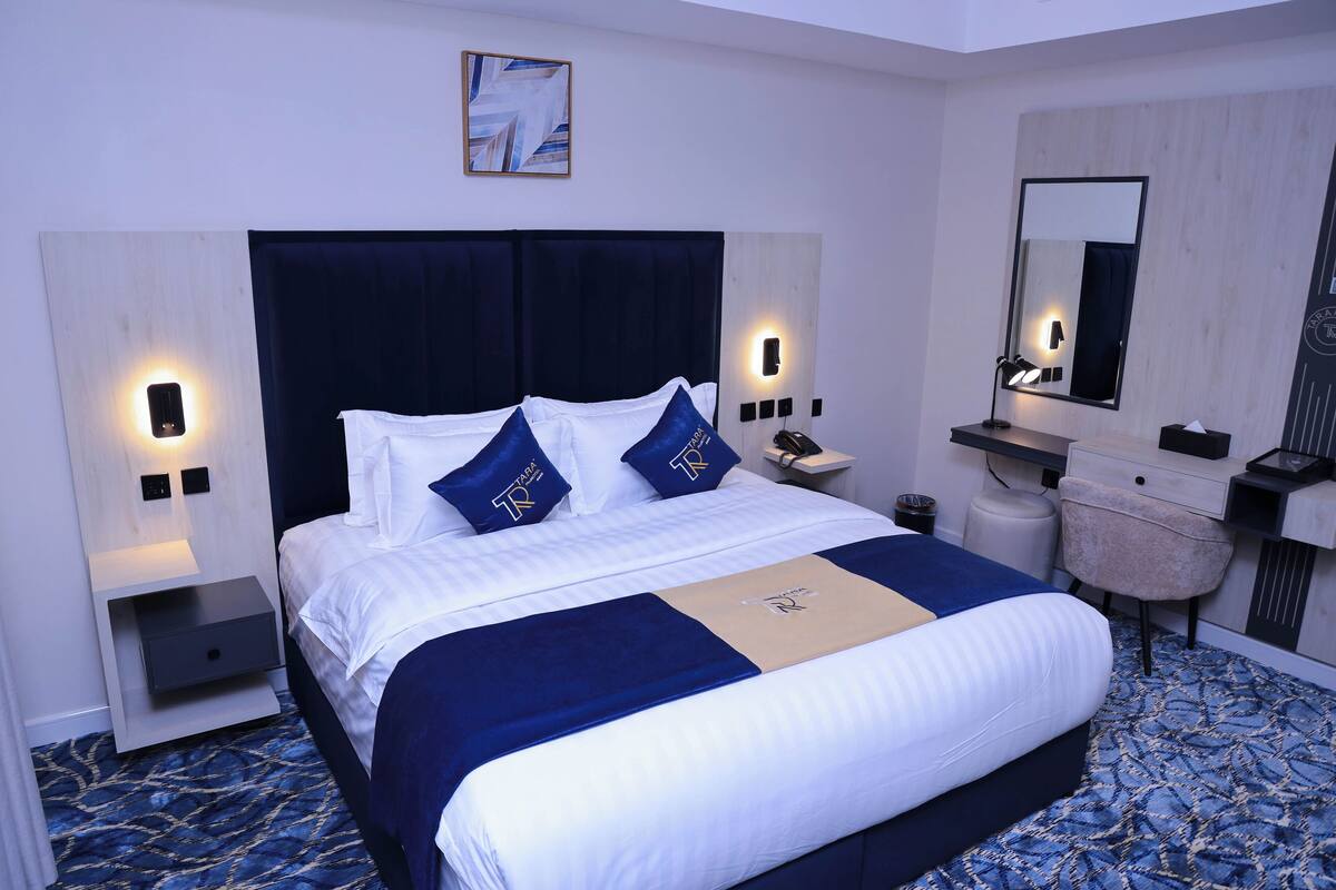 A neatly made bed with a dark headboard, two bedside tables with lamps, a small stool, and a mirror on the wall.