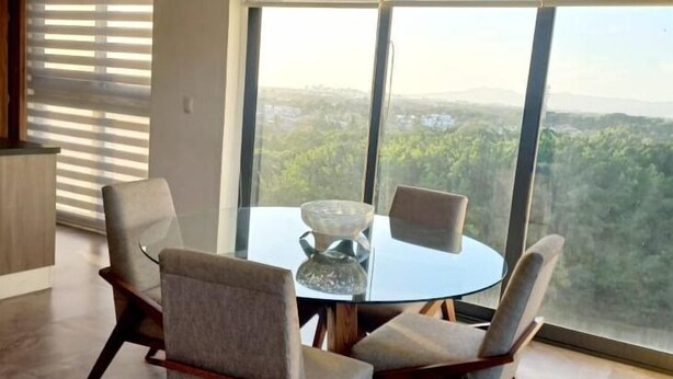 A modern dining area with a round glass table and four chairs, large windows with blinds, and a view of a forested landscape.