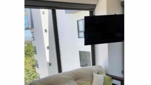 A living room with a sofa, a flat-screen TV, and a window with a view of buildings.
