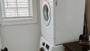 Une buanderie équipée d’un lave-linge et d’un sèche-linge superposés, d’une fenêtre avec des stores et d’un plan de travail de couleur sombre.