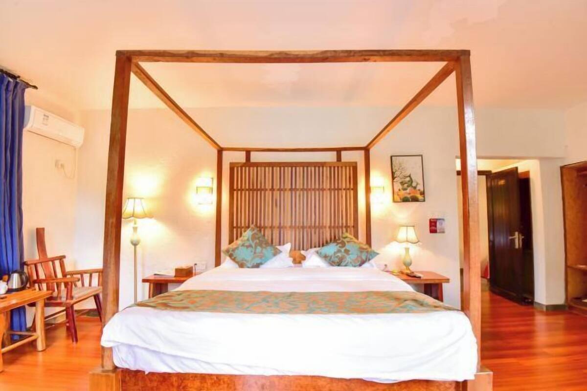 Una cama con dosel de madera, ropa de cama blanca y almohadas decorativas. Hay dos mesitas de noche con lámparas, una silla de madera y un escritorio con silla. El cuarto tiene suelo de madera y una puerta de madera al fondo.