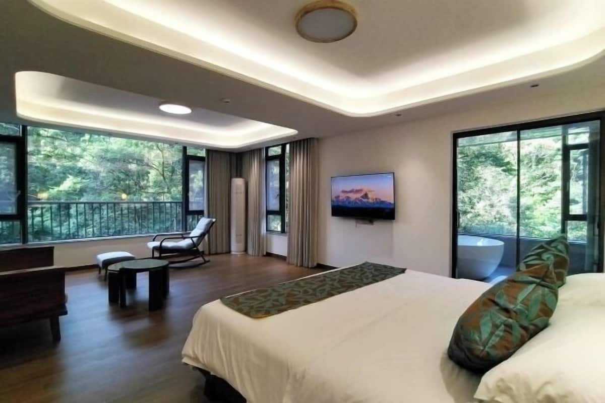 Una habitación de hotel moderna con una cama grande, una zona de estar con una silla y una mesa, y un balcón con vistas a los árboles.