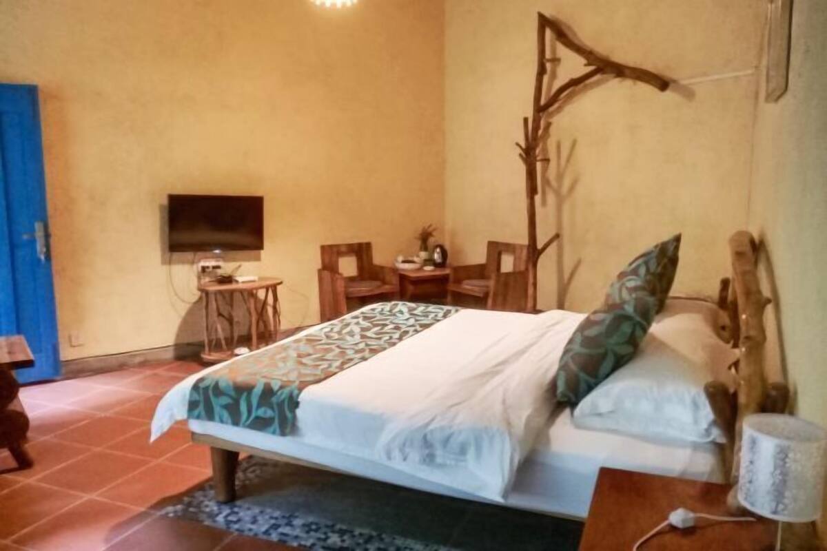 Un dormitorio con una cama, televisor, una mesita, sillas y una rama decorativa en la pared.