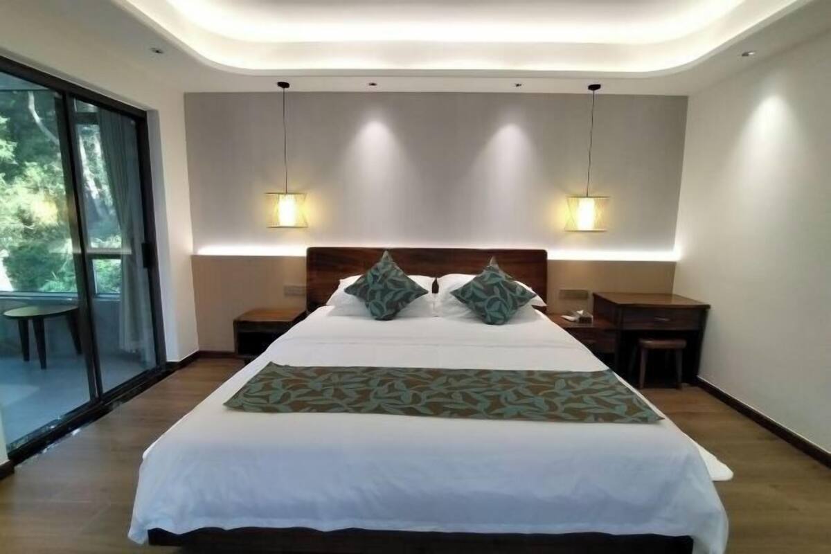 Una habitación de hotel moderna con una cama grande, dos mesitas de noche, un escritorio y una puerta corrediza de vidrio hacia un balcón.
