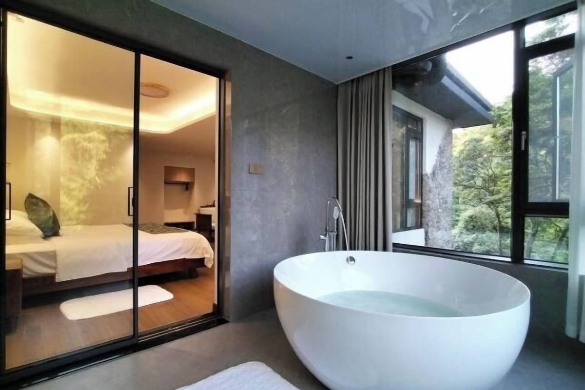 Un baño moderno con una bañera independiente grande, un dormitorio con cama y paredes de vidrio, y vistas a zonas verdes.
