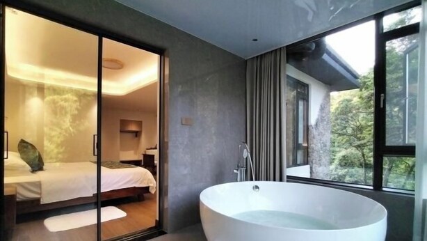 Une salle de bain moderne avec une grande baignoire sur pieds, une chambre fermée par des parois de verre avec un lit, et une vue sur la verdure à l’extérieur.