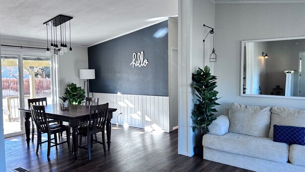 Una sala de estar moderna con piso de madera oscura, un sofá blanco con un cojín azul, un comedor con mesa y sillas de madera oscura, y un espejo grande en la pared.