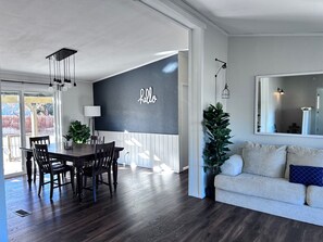 Una sala de estar moderna con piso de madera oscura, un sofá blanco con un cojín azul, un comedor con mesa y sillas de madera oscura, y un espejo grande en la pared.