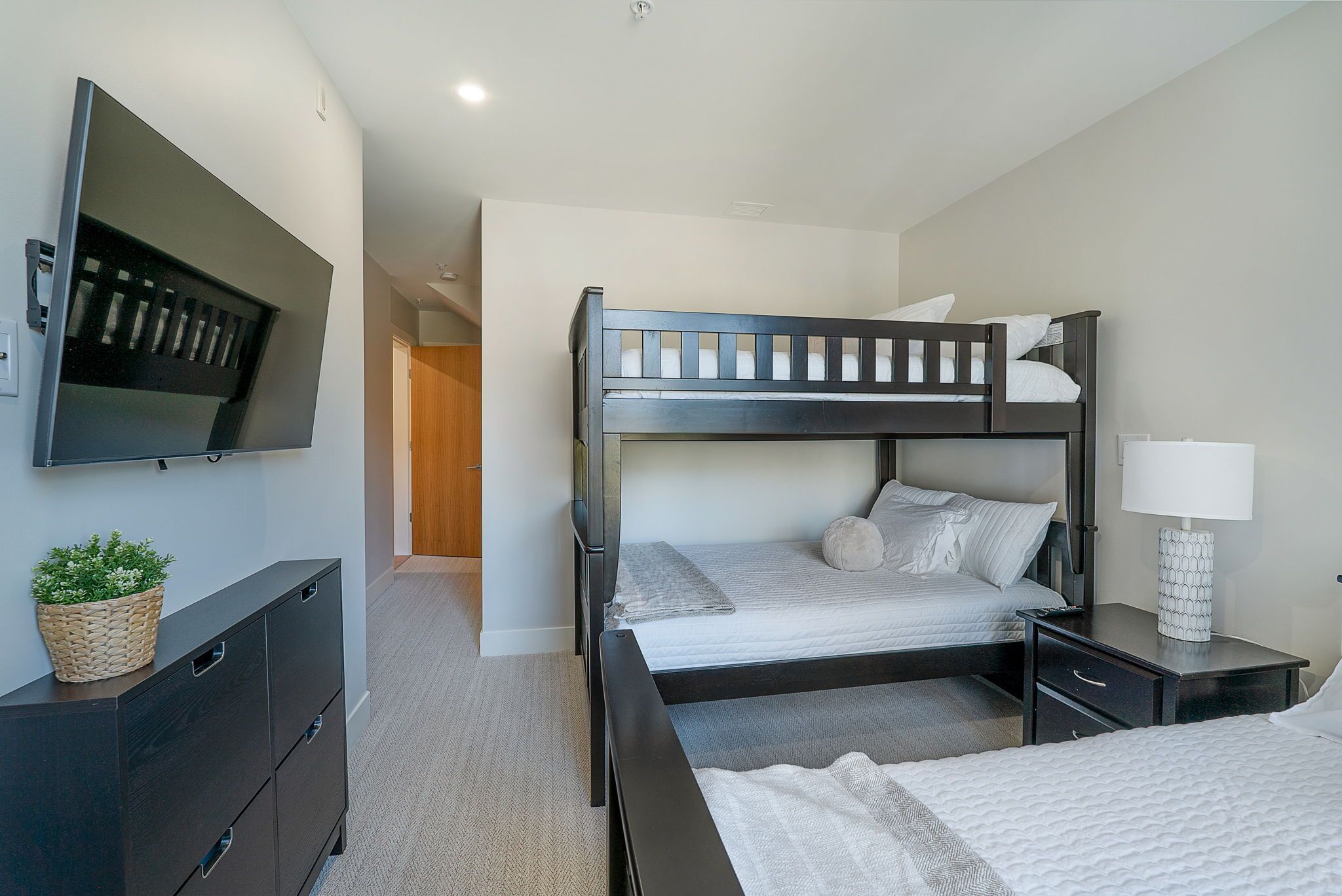A hotel room with a bunk bed, a TV mounted on the wall, a bedside table with a lamp, and a dresser with a plant.
