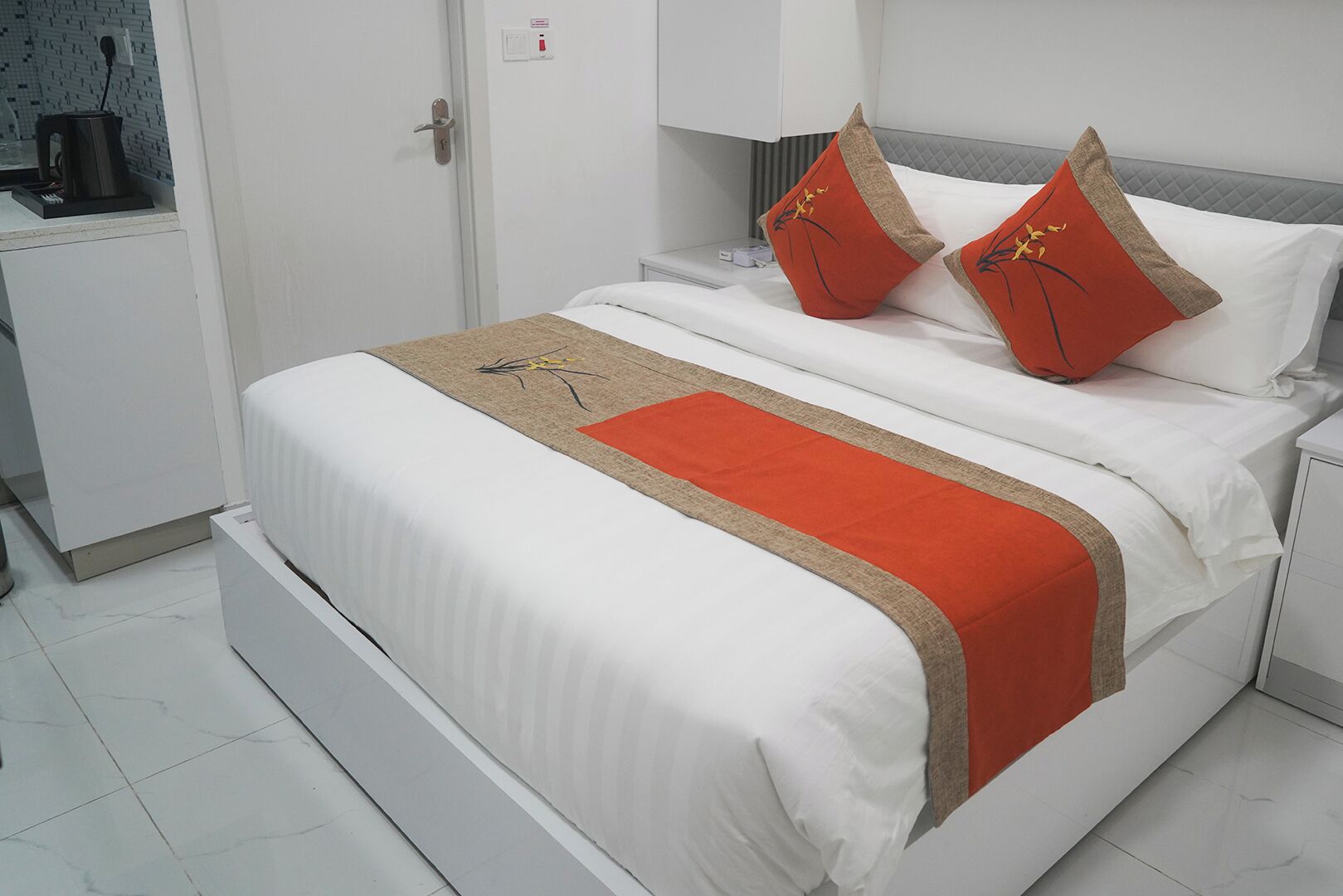 Una cama bien hecha con ropa blanca y dos almohadas rojas, una manta roja y una mesita de noche con una lámpara.