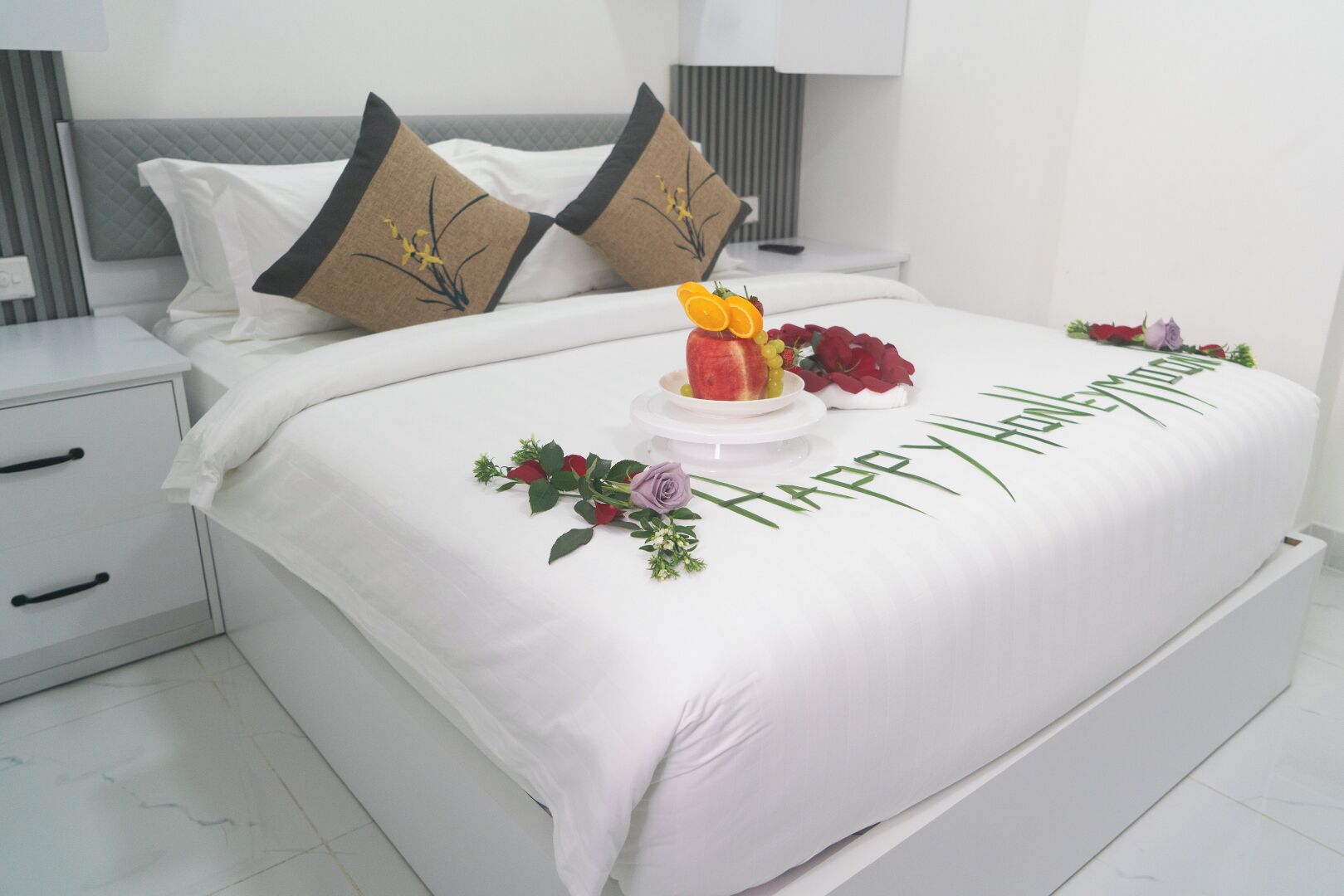 Un lit bien fait, recouvert d’une courtepointe blanche, agrémenté de deux coussins décoratifs, d’une composition florale de fruits et d’un message « Joyeux anniversaire ».