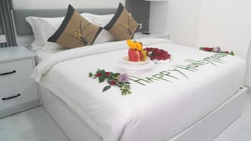 Un lit bien fait, recouvert d’une courtepointe blanche, agrémenté de deux coussins décoratifs, d’une composition florale de fruits et d’un message « Joyeux anniversaire ».