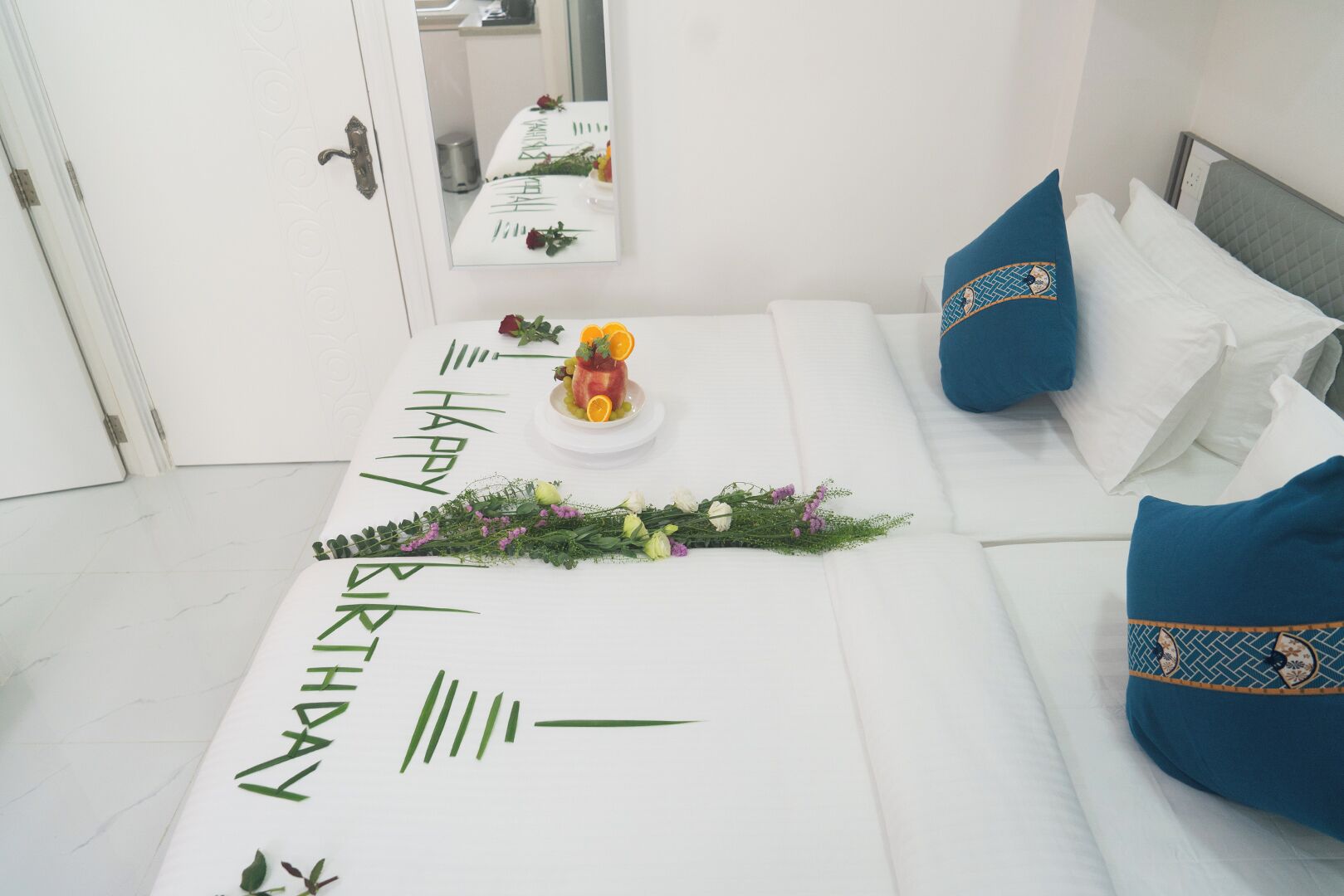 A neatly arranged room with a bed, a small table with a birthday cake, and a mirror reflecting the room.