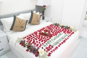 Una cama bien hecha con un mensaje de 'Feliz aniversario' y pétalos de rosa, un jarrón con flores y una mesita de noche con una lámpara.