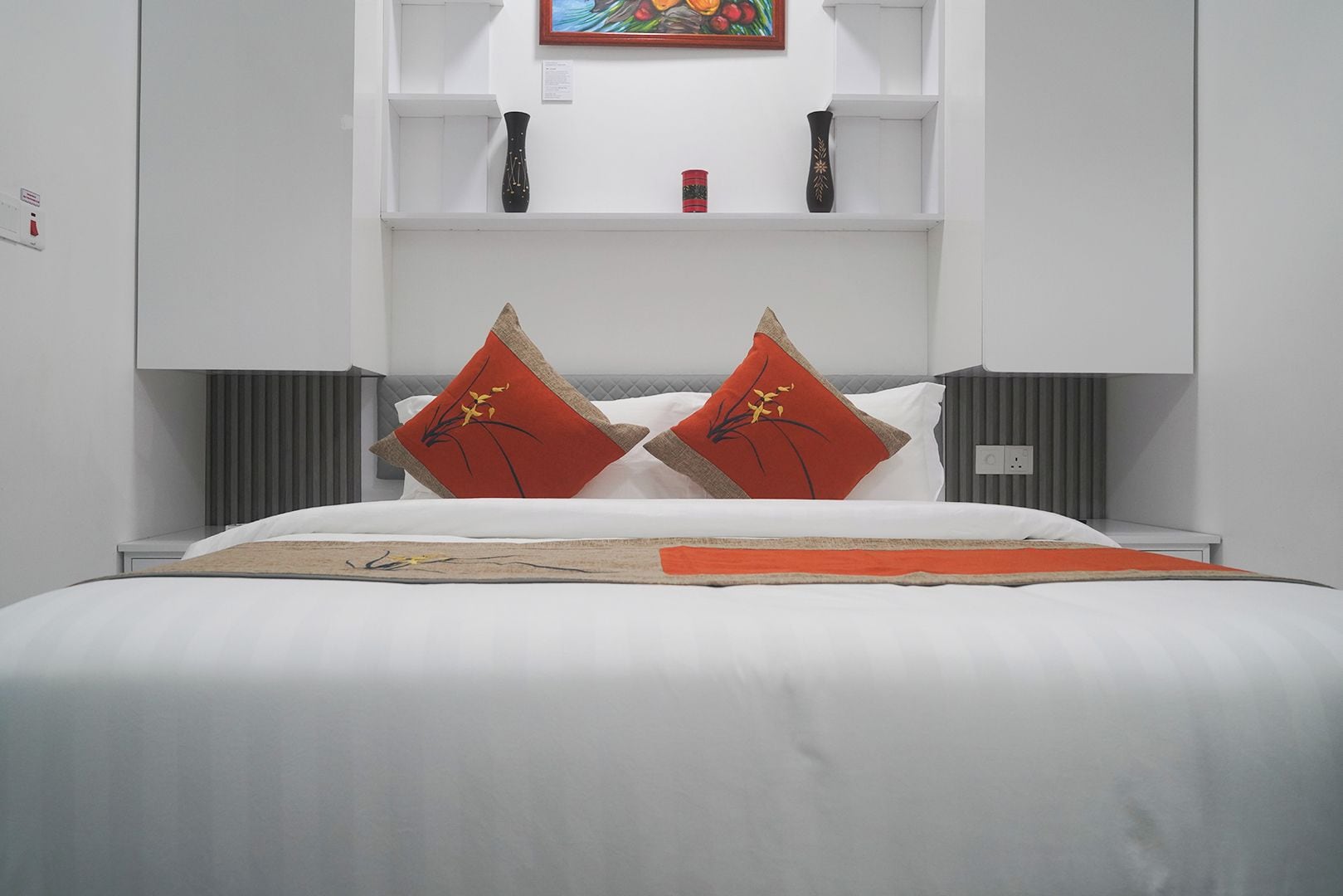 A modern hotel room with a large bed, two red decorative pillows, a white headboard, and a shelf with a vase and a small painting.