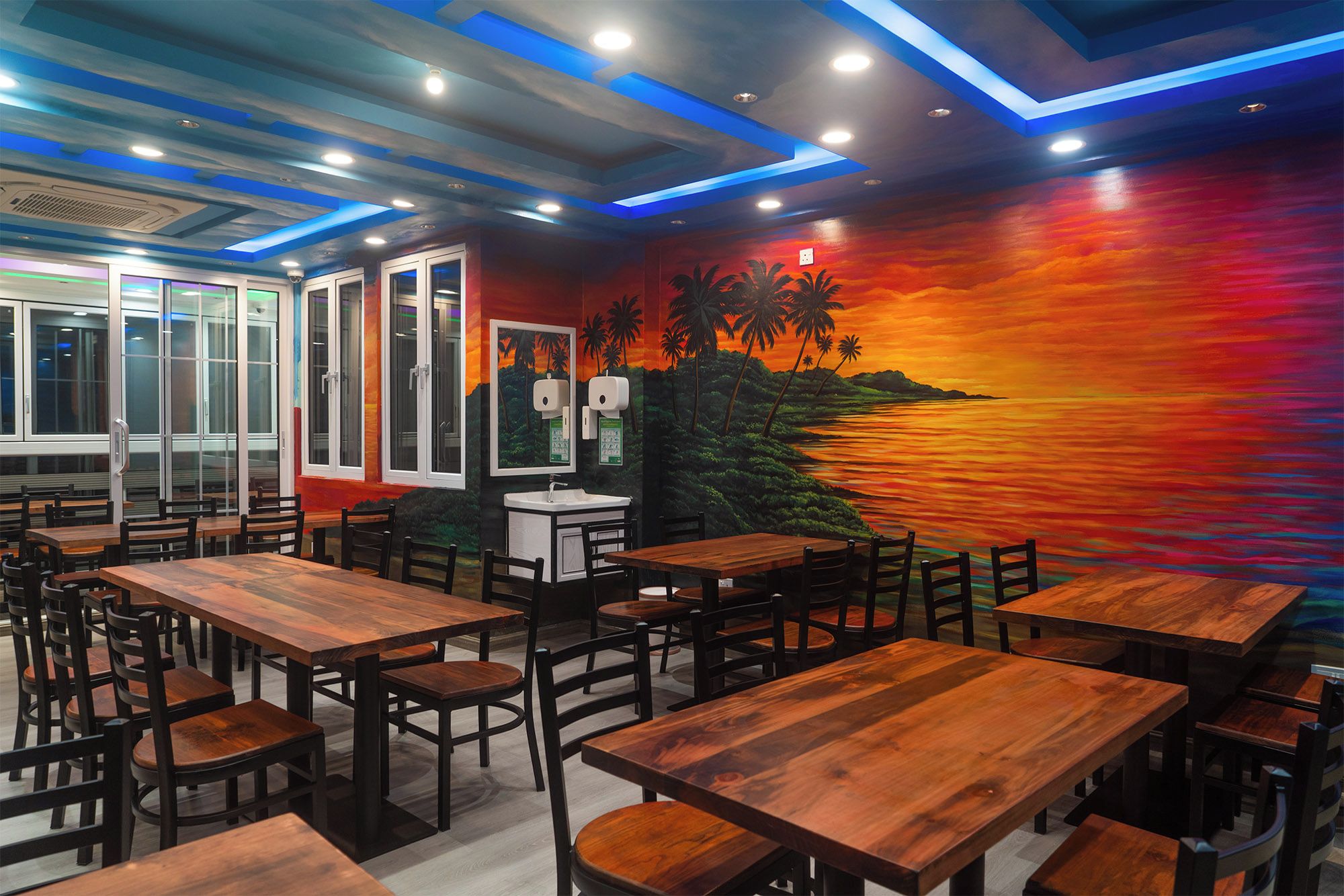 A restaurant interior with wooden tables and chairs, a mural of a sunset, and a view of the sea.
