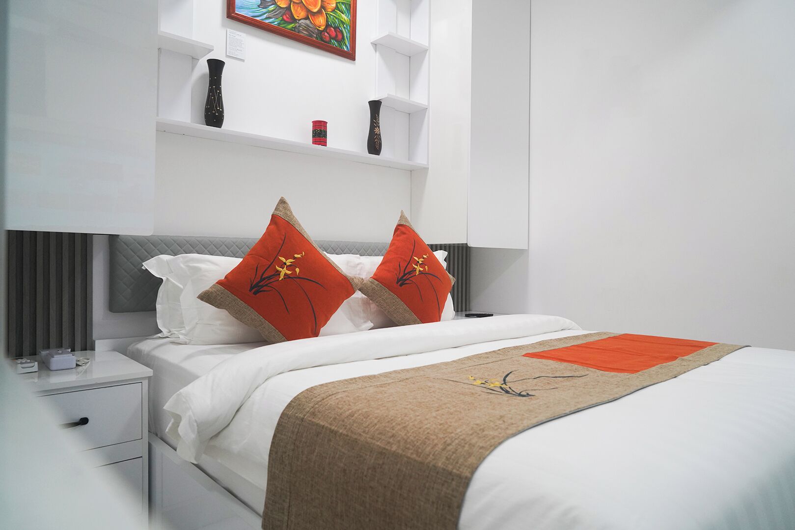 A modern hotel room with a neatly made bed, a bedside table with a lamp, and a shelf with decorative items.