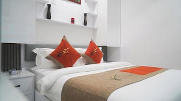 A modern hotel room with a neatly made bed, a bedside table with a lamp, and a shelf with decorative items.