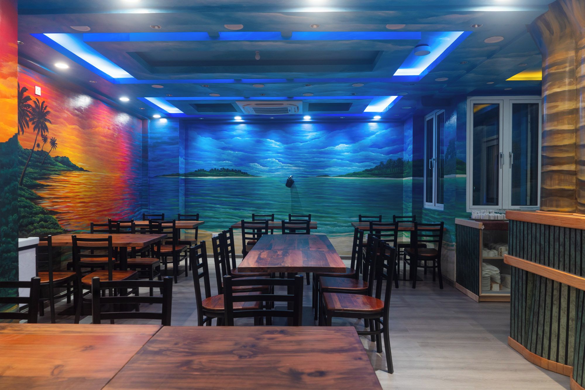 Een restaurant met een muurschildering van een zonsondergang boven het water, houten tafels en stoelen, en uitzicht op de zee door grote ramen.
