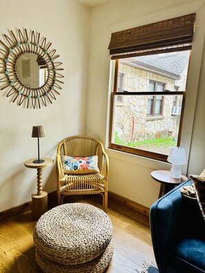 A room with a wicker chair, a round woven ottoman, a small table with a lamp, and a large window with a view of a stone building. - Casita Rosedale - Central Austin (Austin)