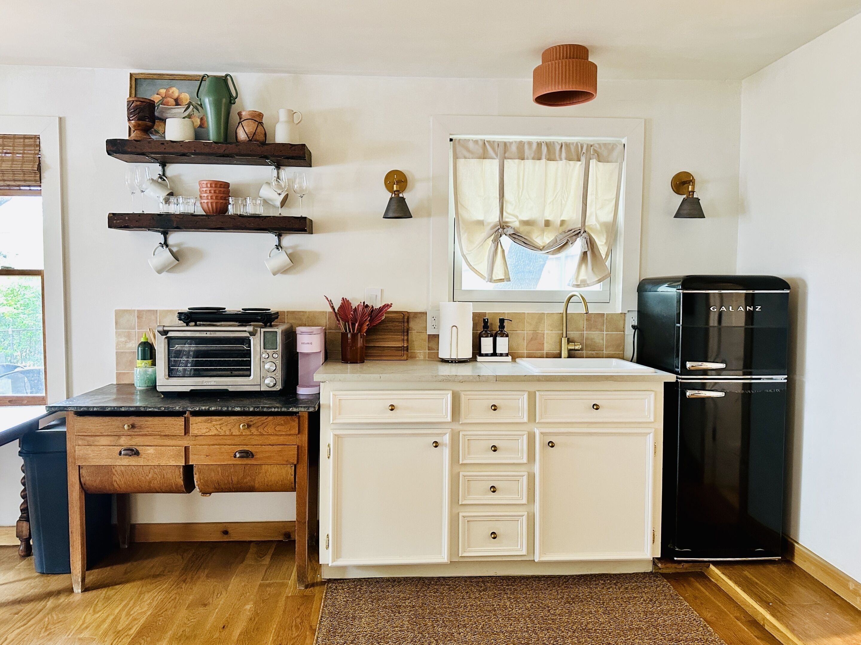 A kitchen with a microwave, toaster, sink, and black refrigerator. There are wooden cabinets, a window with curtains, and a shelf with decorative items.