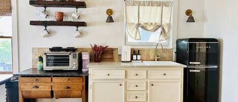 A kitchen with a microwave, toaster, sink, and black refrigerator. There are wooden cabinets, a window with curtains, and a shelf with decorative items.
