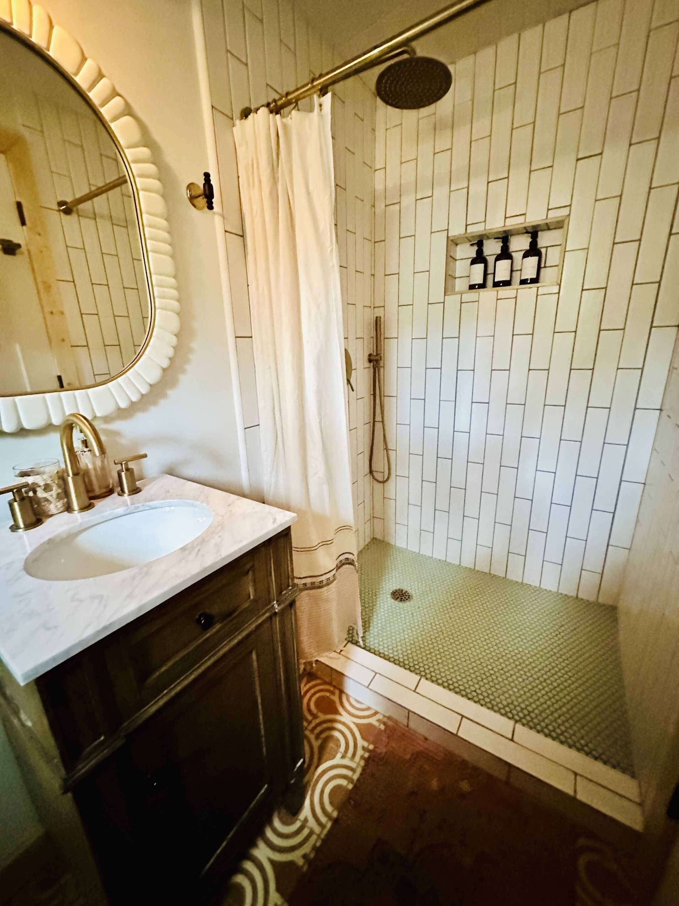 A bathroom with a white tiled shower, a sink with a marble countertop, and a mirror with a scalloped edge.