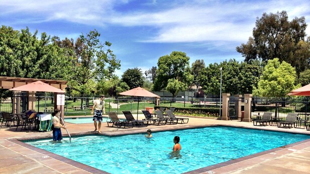 Una piscina con sillas de descanso y sombrillas, rodeada de árboles y una zona vallada.