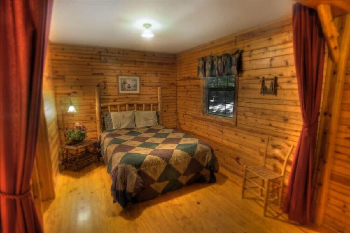 A cozy log cabin bedroom with a checkered bedspread, a wooden headboard, a small bedside table with a lamp, and a window with blinds.