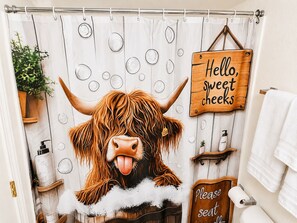 A bathroom with a shower curtain featuring a bull's face, a 'Hello, sweet cheeks' sign, and a 'Please seat yourself' sign.