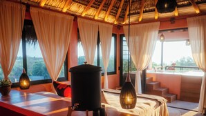 A room with a wooden ceiling, large windows with white curtains, a bed with a white bedspread, a black trash bin, and a small table with a plant and a lamp. - Penthouse with pool & ocean view (Puerto Escondido)