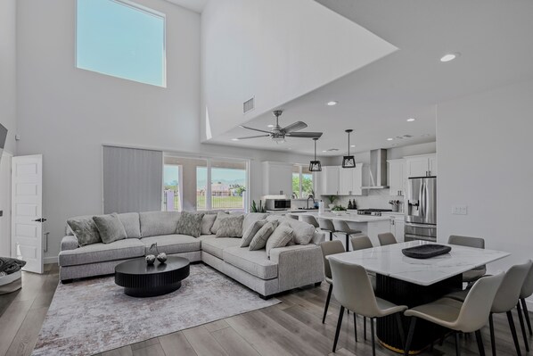 A modern living room with a sectional sofa, a dining table with chairs, a ceiling fan, and a kitchen area with cabinets and appliances.