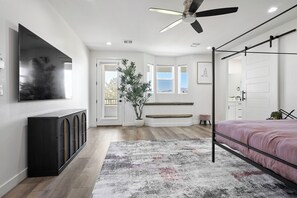 A bedroom with a bed, a TV, a ceiling fan, a window, and a plant.