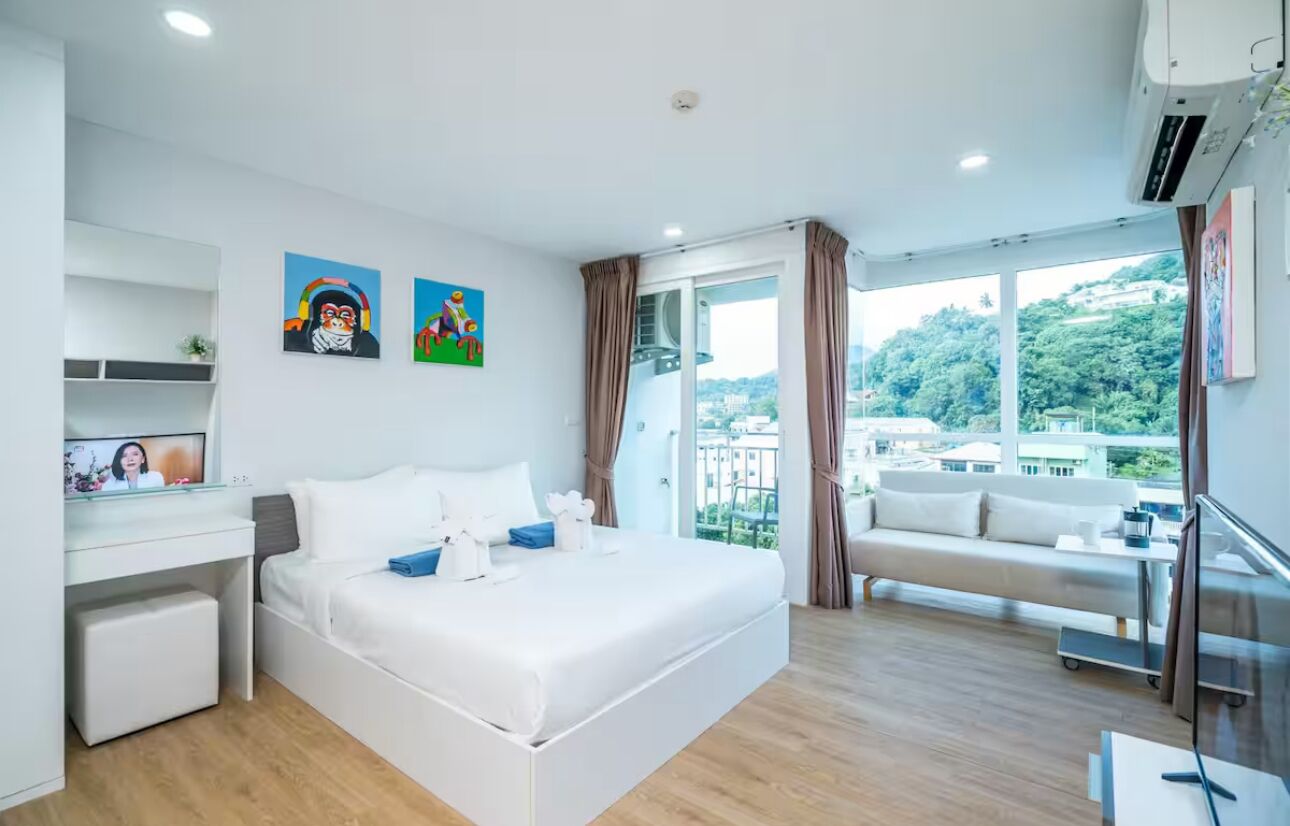 A modern hotel room with a large bed, a desk, and a balcony with a city view.