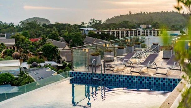Une piscine sur le toit, dotée de chaises longues, d’une balustrade en verre et offrant une vue sur les bâtiments et la végétation environnants.