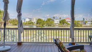 A covered outdoor area with a wooden deck, a cushioned bench, and a view of a lake and houses in the background. - Marina & Golf Course Views: Edisto Island Home (Edisto Island)