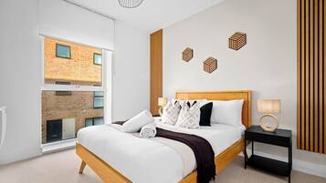 A modern bedroom with a large bed, a nightstand, a lamp, and a view of a brick building through a large window.