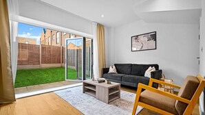 A modern living room with a sofa, armchair, and coffee table. A sliding glass door leads to a backyard with a lawn and a fence. - Luxury 4 Bedroom Townhouse - Garden - Balconies (Birmingham)