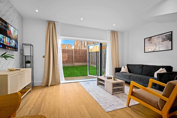 A modern living room with a grey sofa, wooden coffee table, and a small wooden chair. A TV is mounted on the wall, and there is a sliding glass door leading to a backyard. - Stylish 4 Bedroom Townhouse - Garden - Balconies - Parking (Birmingham)