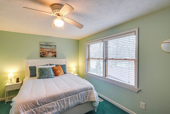 A bedroom with a bed, a ceiling fan, a window with blinds, a nightstand with a lamp, and a painting on the wall. - Lakefront Getaway w/ Fire Pit in Akron (Akron)
